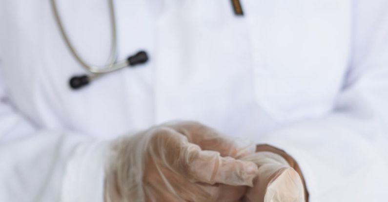 Diagnostic Test - Faceless ethnic medical worker in lab coat and stethoscope taking of transparent gloves after approaching patients for examination in modern hospital