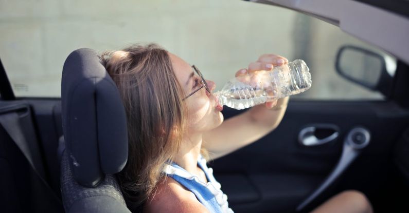 Car Fluids - High angle side view of young woman in glasses and casual clothes drinking water from plastic transparent bottle while sitting in cabriolet with open roof in traffic jam in hot sunny day