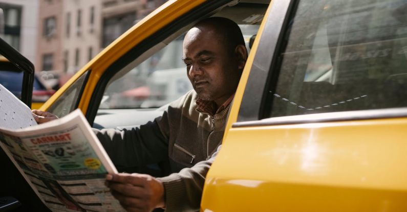 Daily Commute - Concentrated male driver in casual clothes sitting in taxi during break and reading newspaper